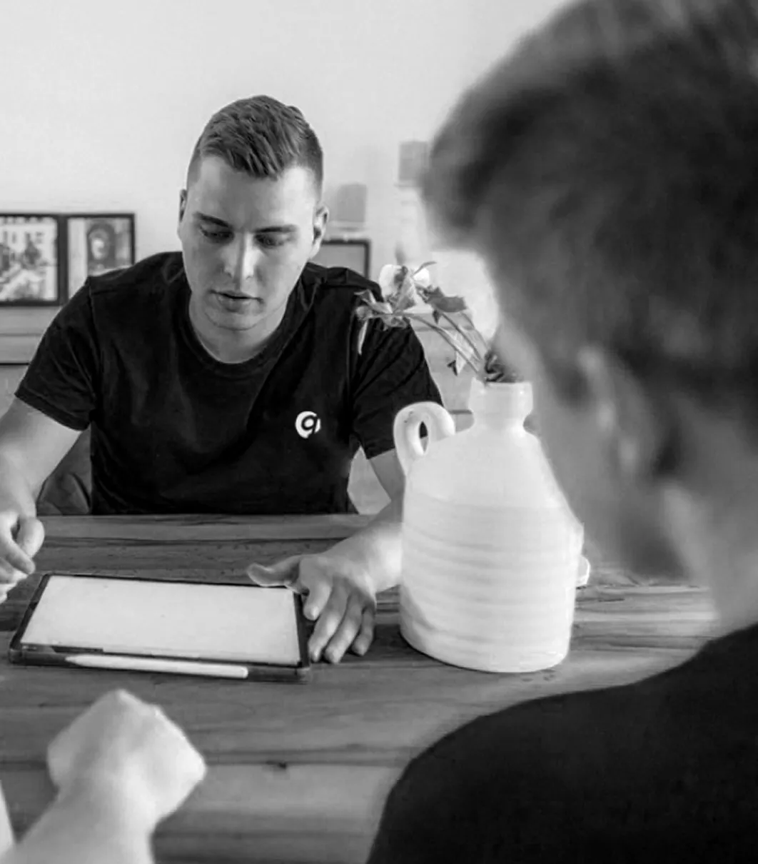 Zwei Männer sitzen an einem Holztisch und besprechen etwas. Einer von ihnen zeigt auf ein Tablet mit Eingabestift. Beide tragen dunkle T-Shirts, einer davon mit einem Logo auf der Brust. Im Hintergrund steht eine Vase mit Blumen und eingerahmte Fotos. Das Bild ist in Schwarz-Weiß gehalten.